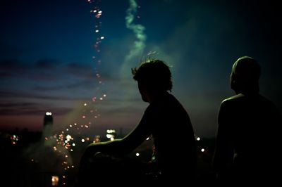 Two people in right foreground watch a stream of fireworks launch in the left background